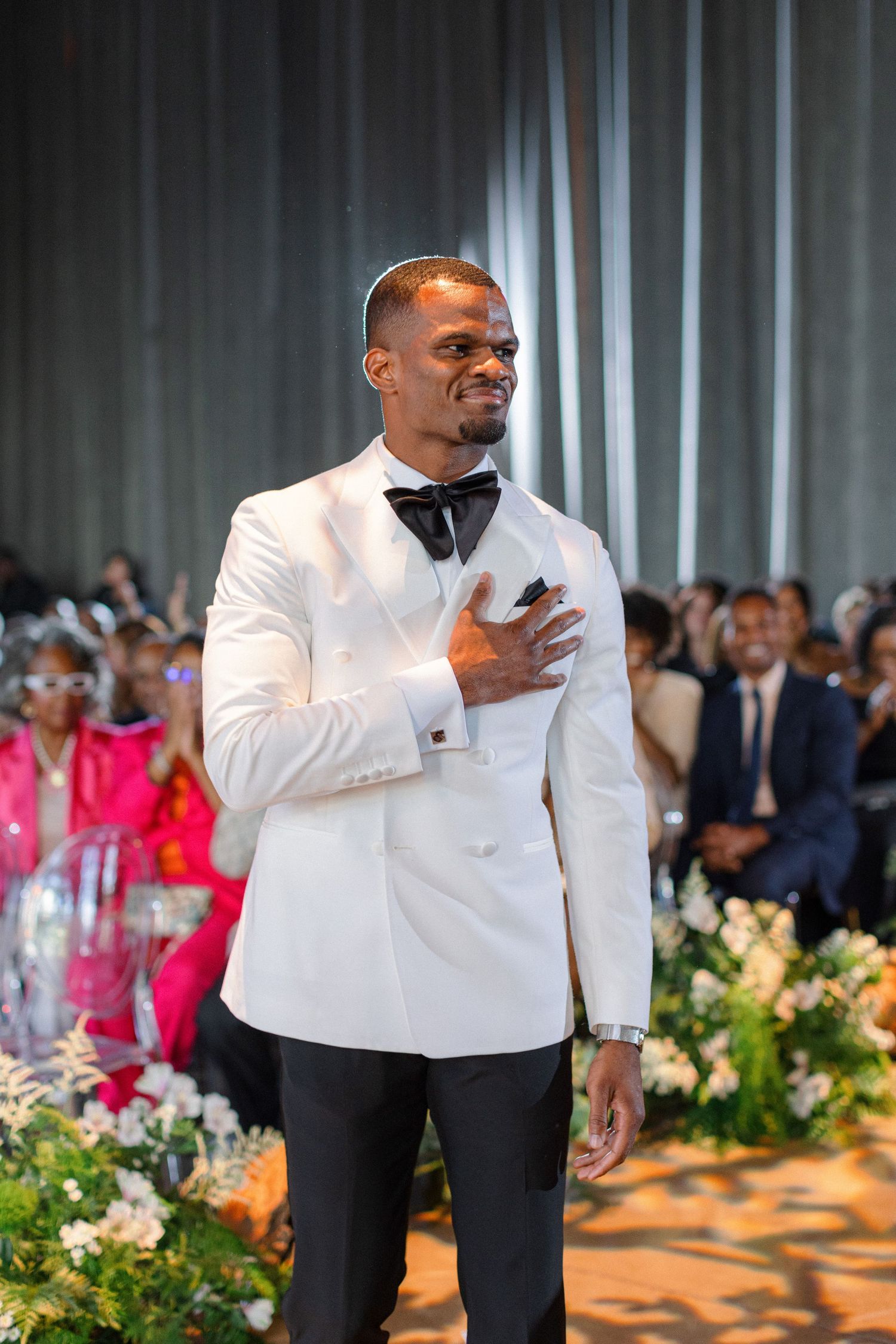Emotional groom in white tuxedo places hand on heart during wedding ceremony.