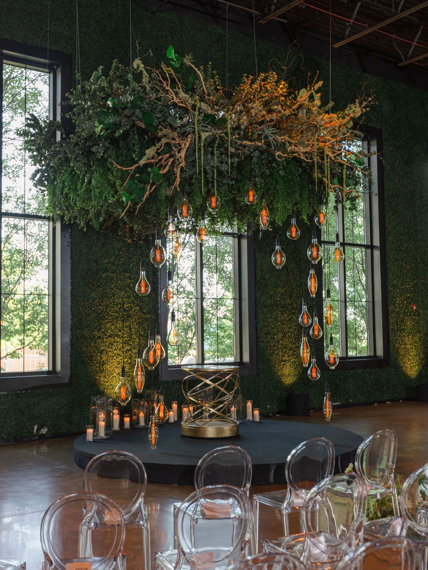 Modern wedding reception venue features hanging greenery and Edison bulb lighting installation above ghost chairs.