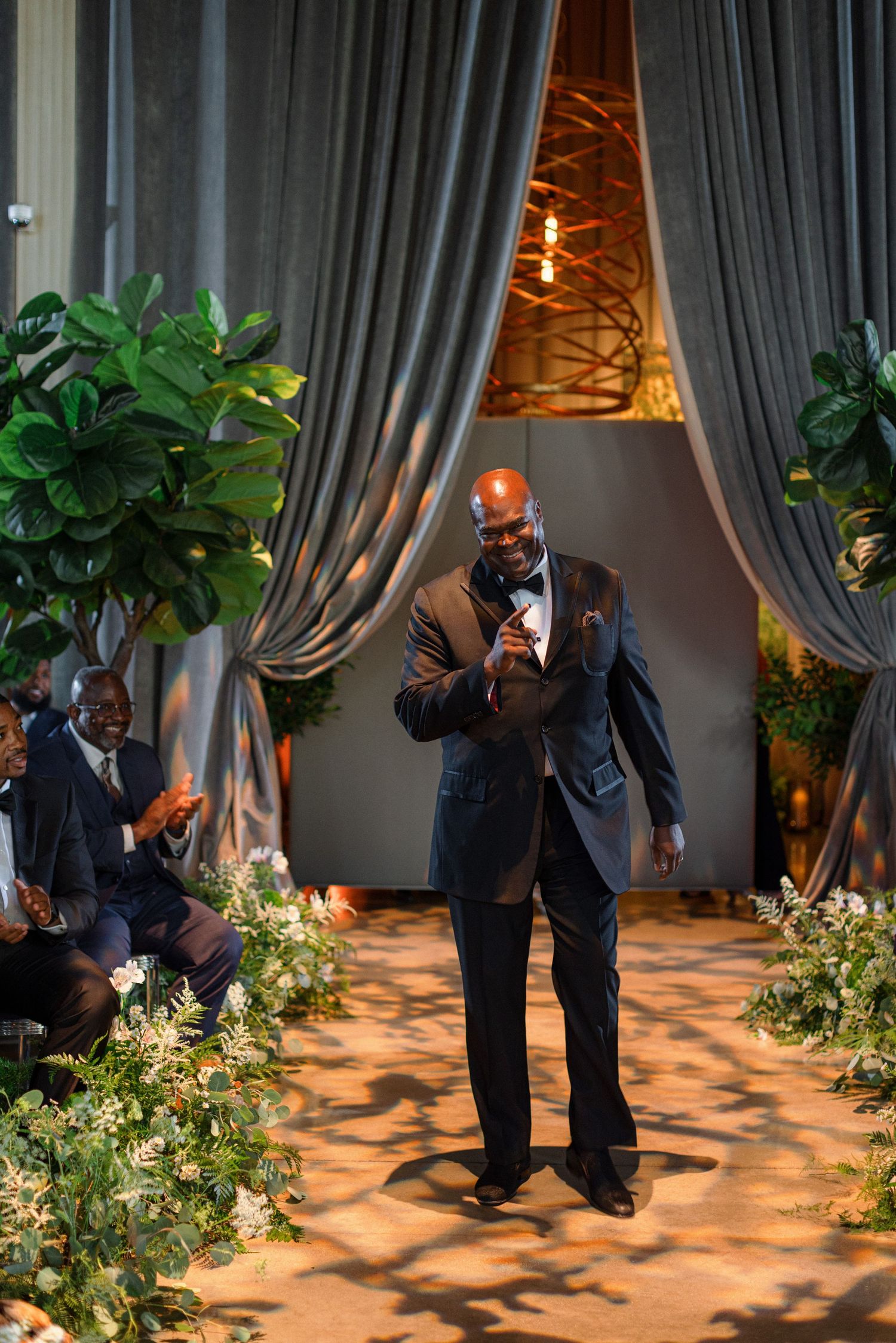 Groom in dark suit delivers speech during wedding reception surrounded by elegant decor.