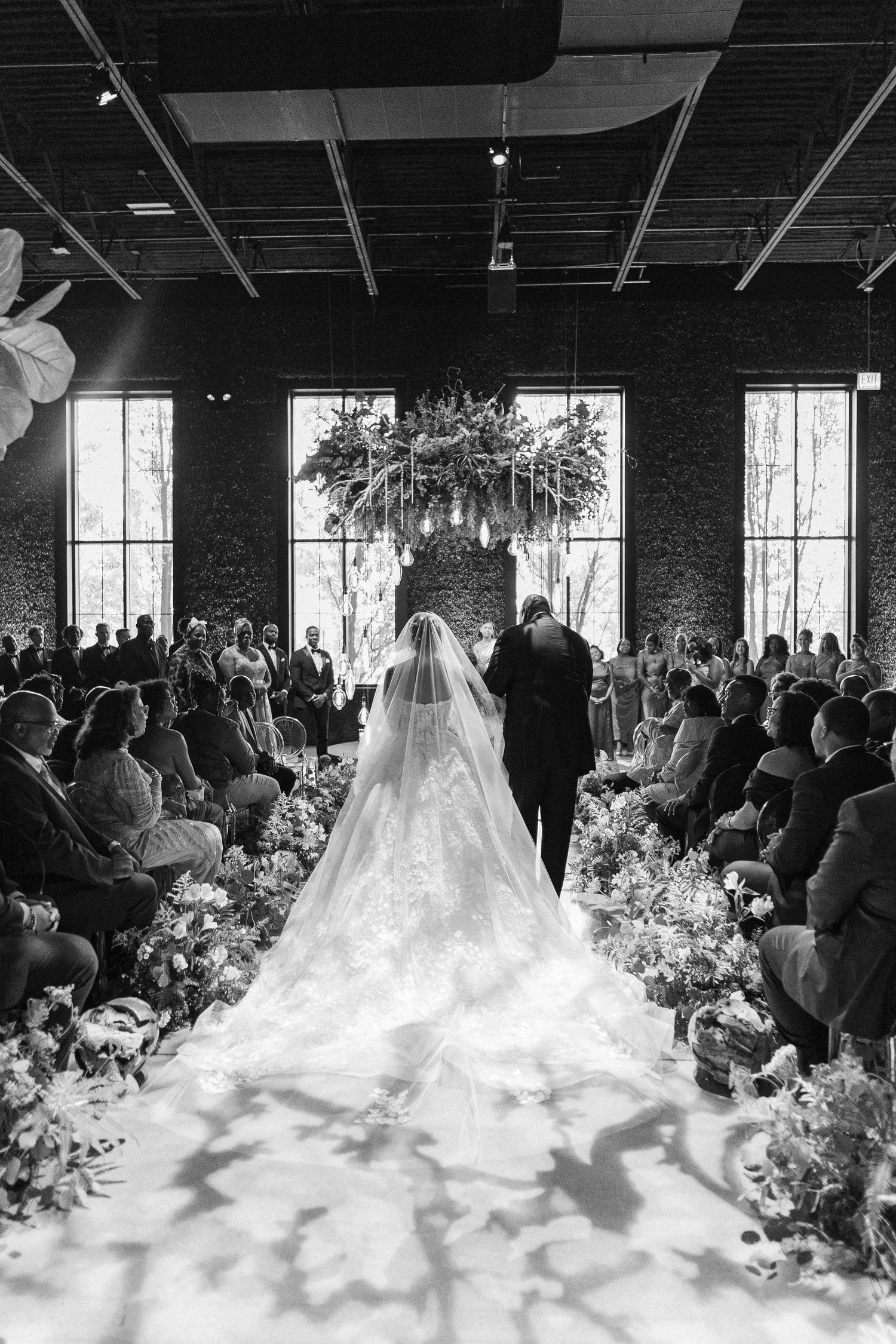 Dramatic black and white photo of bride walking down flower-lined aisle with flowing cathedral veil.