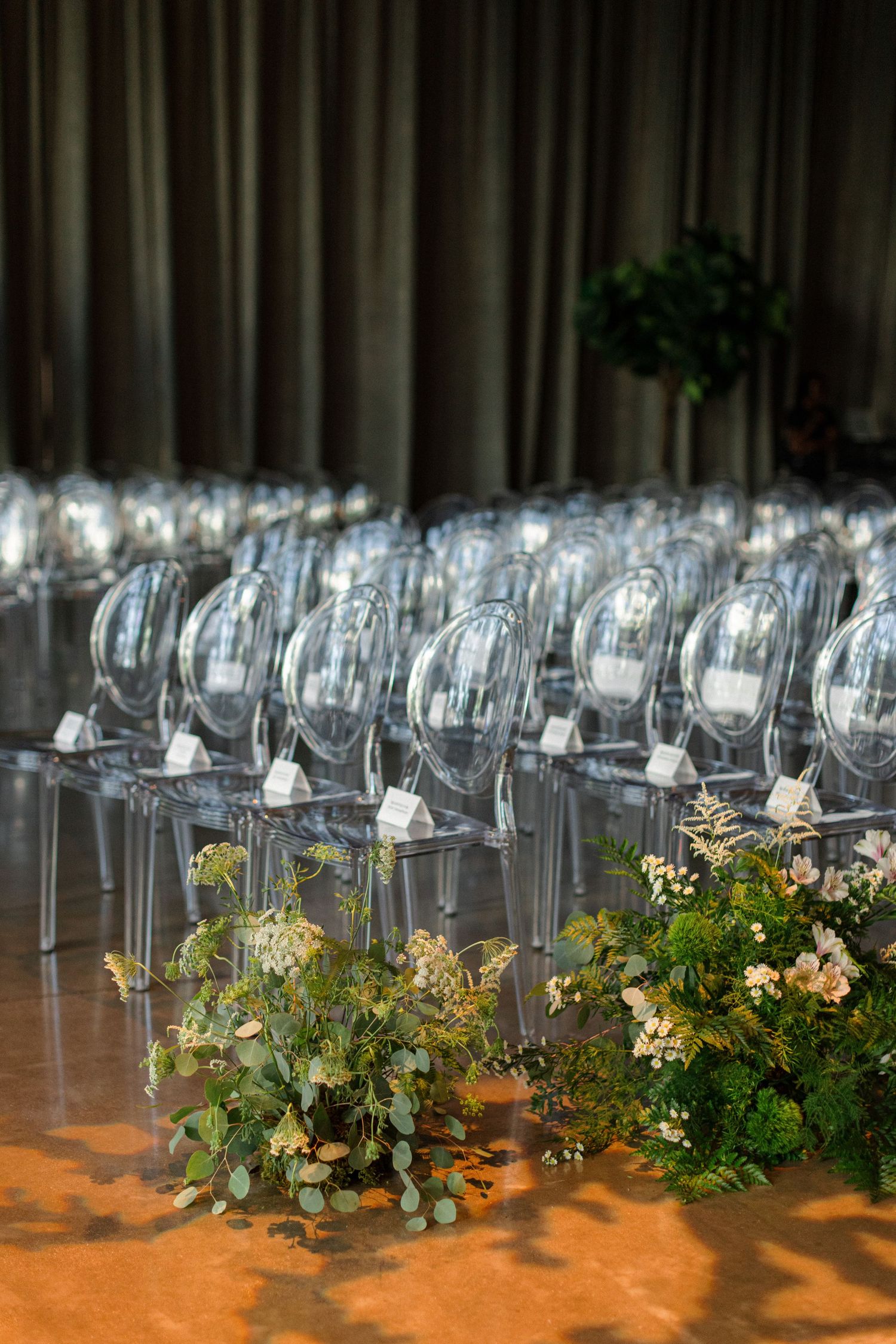 Ghost chairs arranged in rows with delicate floral arrangements and greenery for wedding ceremony seating.