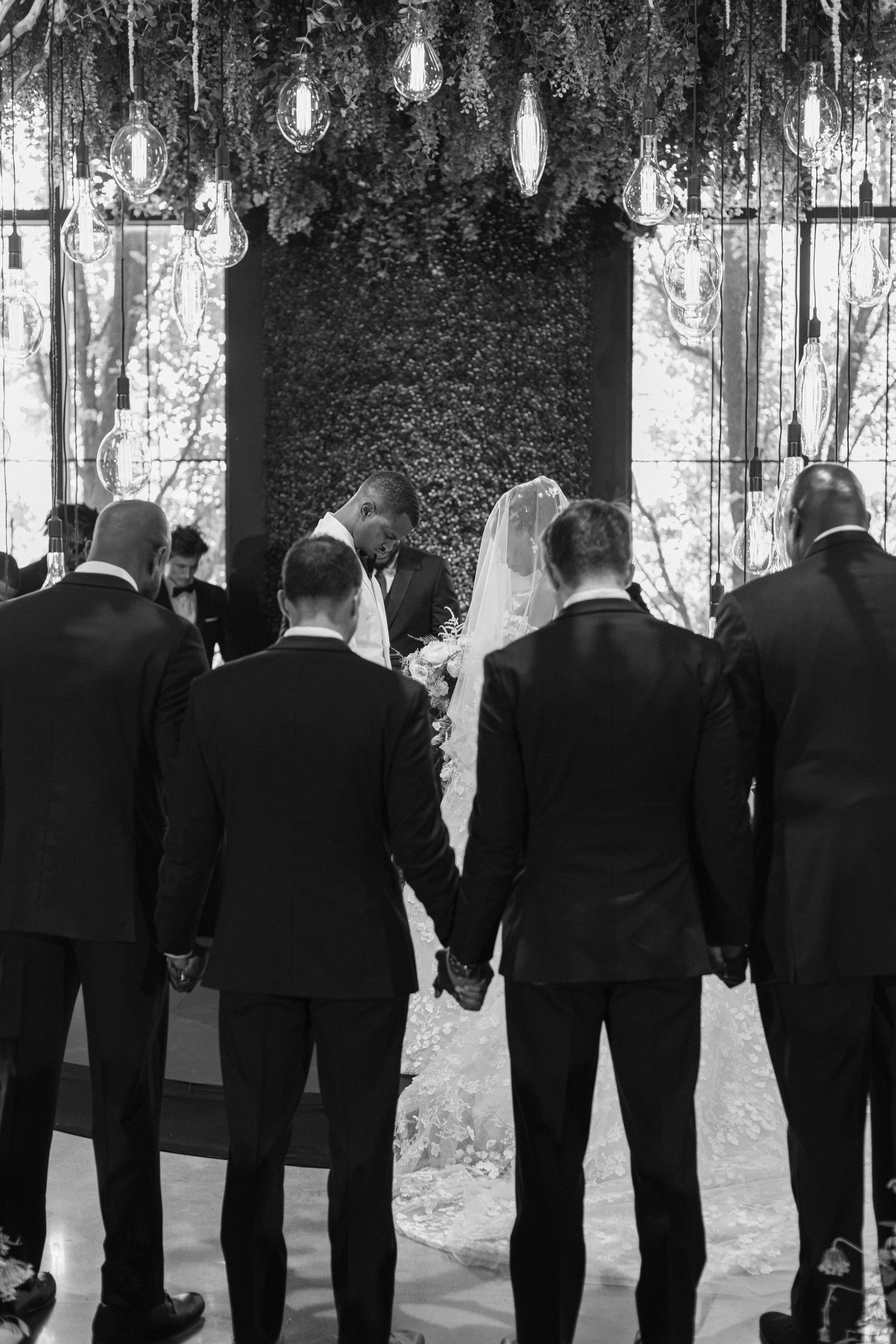 Groomsmen in black suits stand at the altar under hanging lights and greenery during a wedding ceremony.