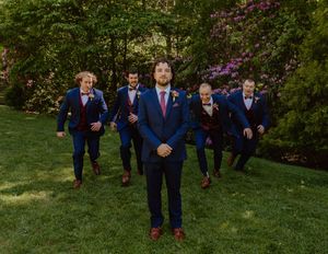 Groomsmen in navy suits pose playfully on green grass with flowering trees in the background.