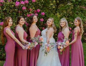 Bridesmaids in mauve dresses stand together with bouquets at an outdoor wedding ceremony.