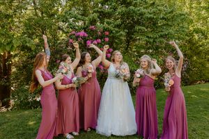 Wedding party in mauve and white dresses celebrates joyfully on green lawn.