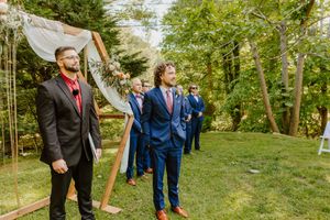 Groomsmen in navy suits and red ties stand under trees at outdoor ceremony.