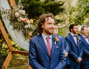 Wedding party stands beneath wooden arch decorated with white fabric and greenery.