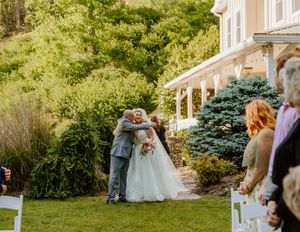 Outdoor wedding ceremony setup with white chairs and wooden arch against house backdrop.