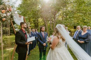 Wedding ceremony guests gathered on white chairs in a sunlit garden setting.