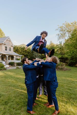 Groomsmen in navy suits performing acrobatic jumps and poses on grass.