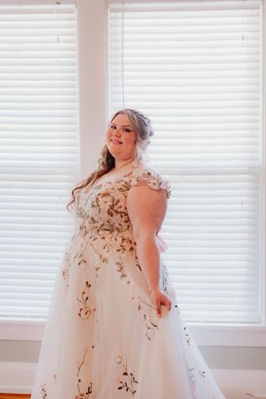A bride poses in a white floral embellished wedding gown against a window backdrop.