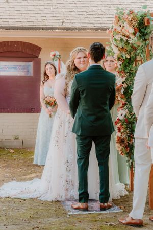 Wedding party members stand together against a white brick wall with floral decorations.