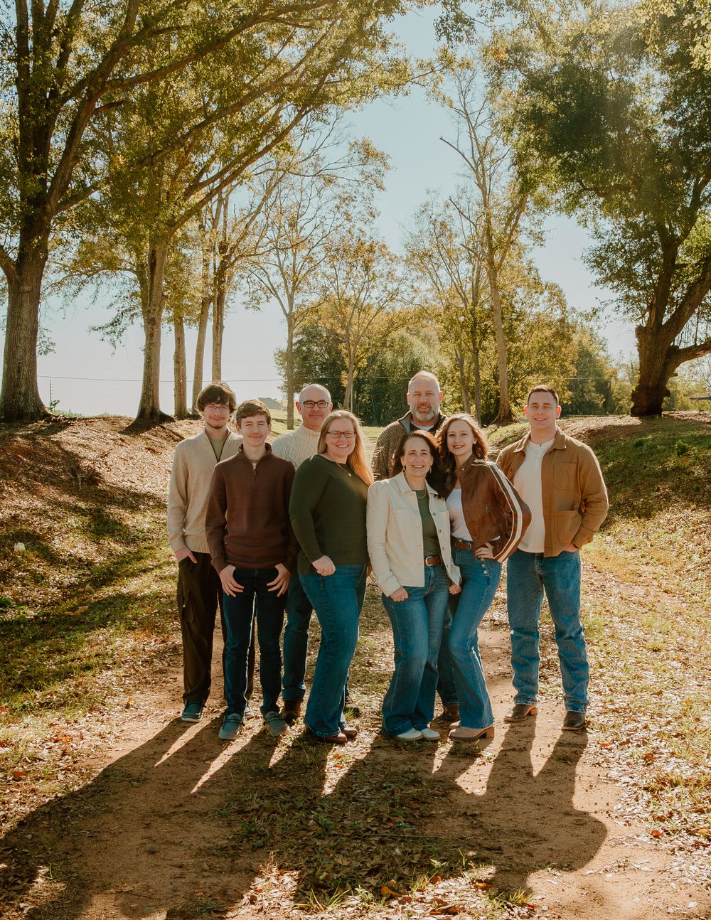 Extended family poses together on a fall day with golden sunlight filtering through trees.