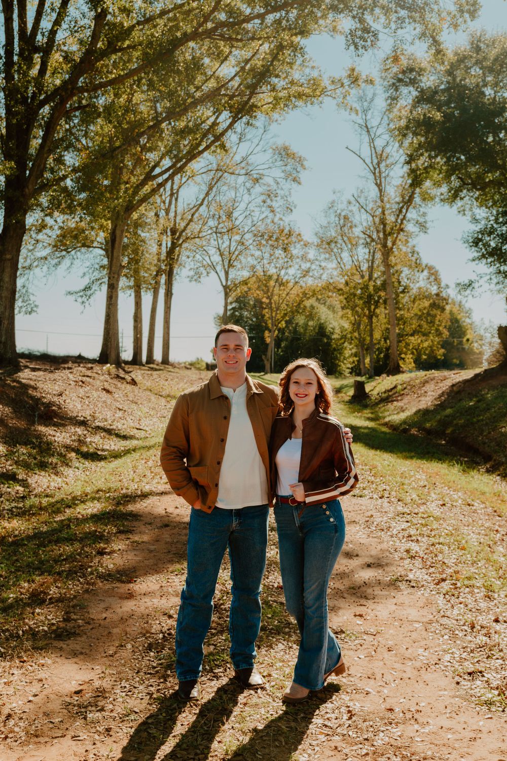 Couple standing together on a sunlit autumn path surrounded by tall trees.