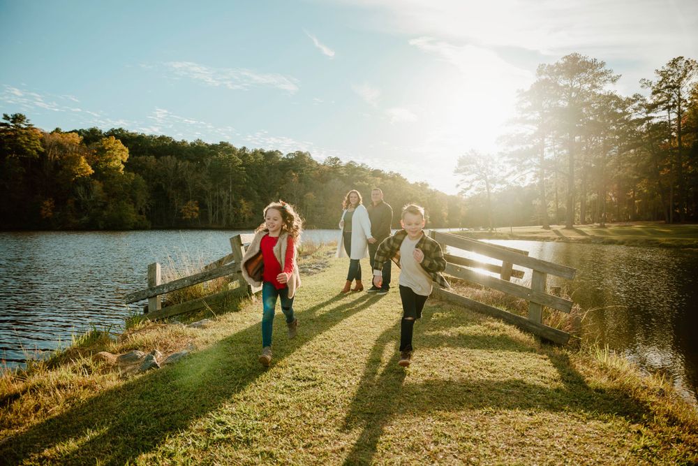 People walking along a grassy lakeside path with sunlight streaming through pine trees.