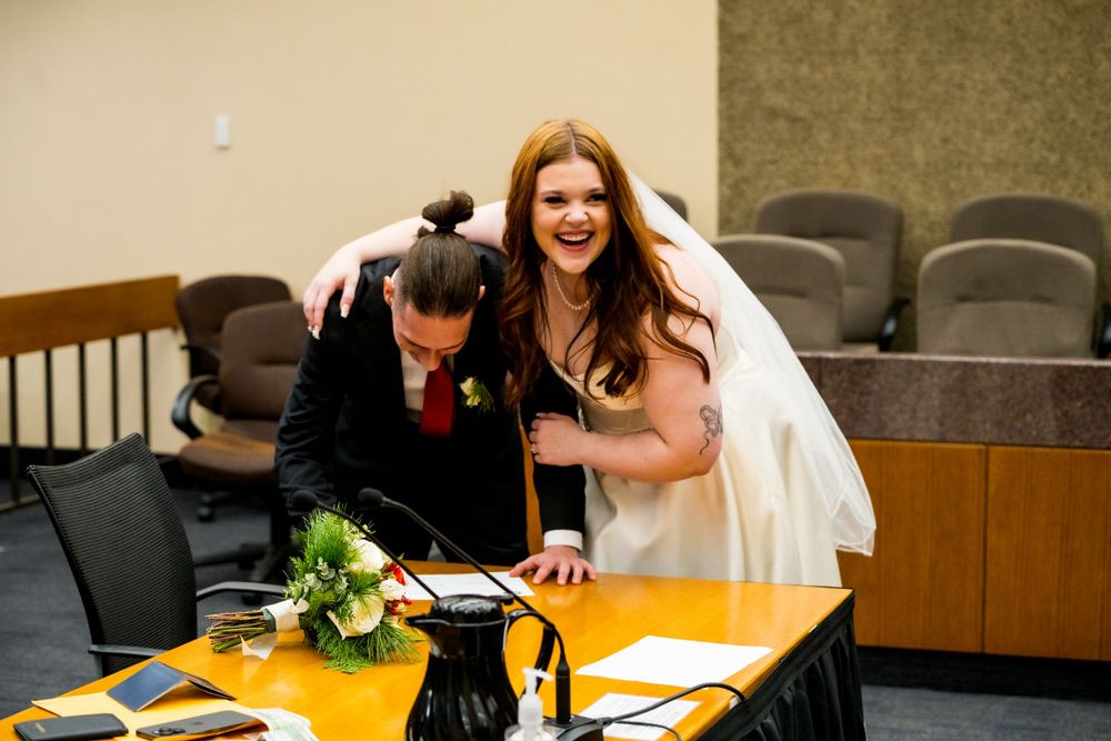 Two people laughing joyfully while signing marriage paperwork at a formal desk.
