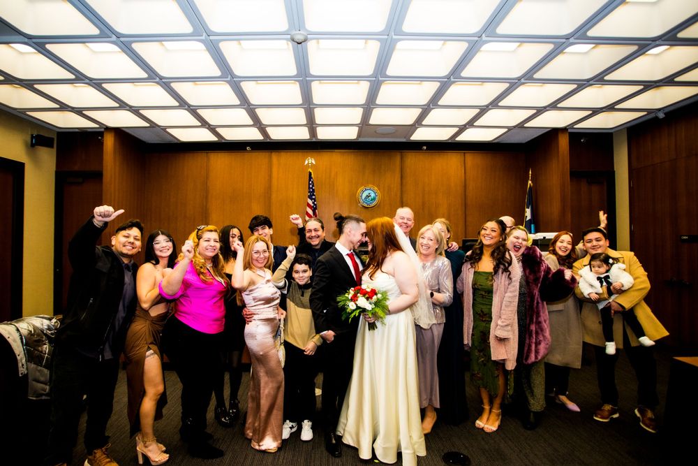 Group photo of wedding party celebrating with raised arms under geometric ceiling lighting.