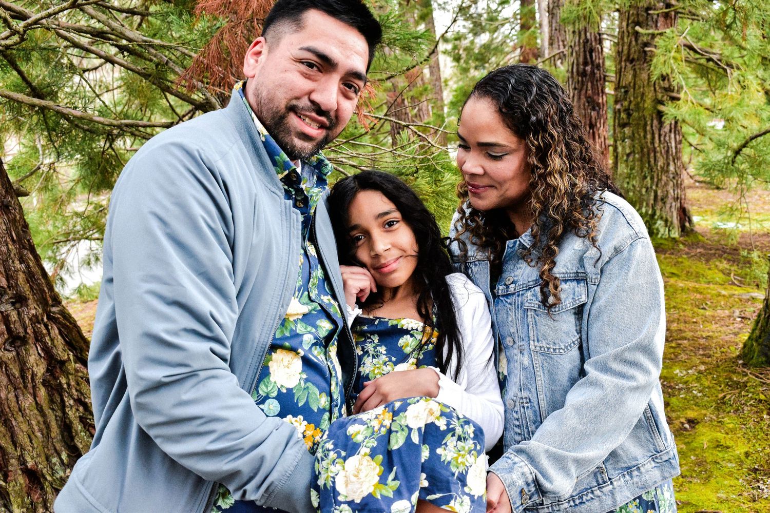 Seattle family photography. Family in coordinating blue and grey outfits stand close together while posing outdoors among trees.