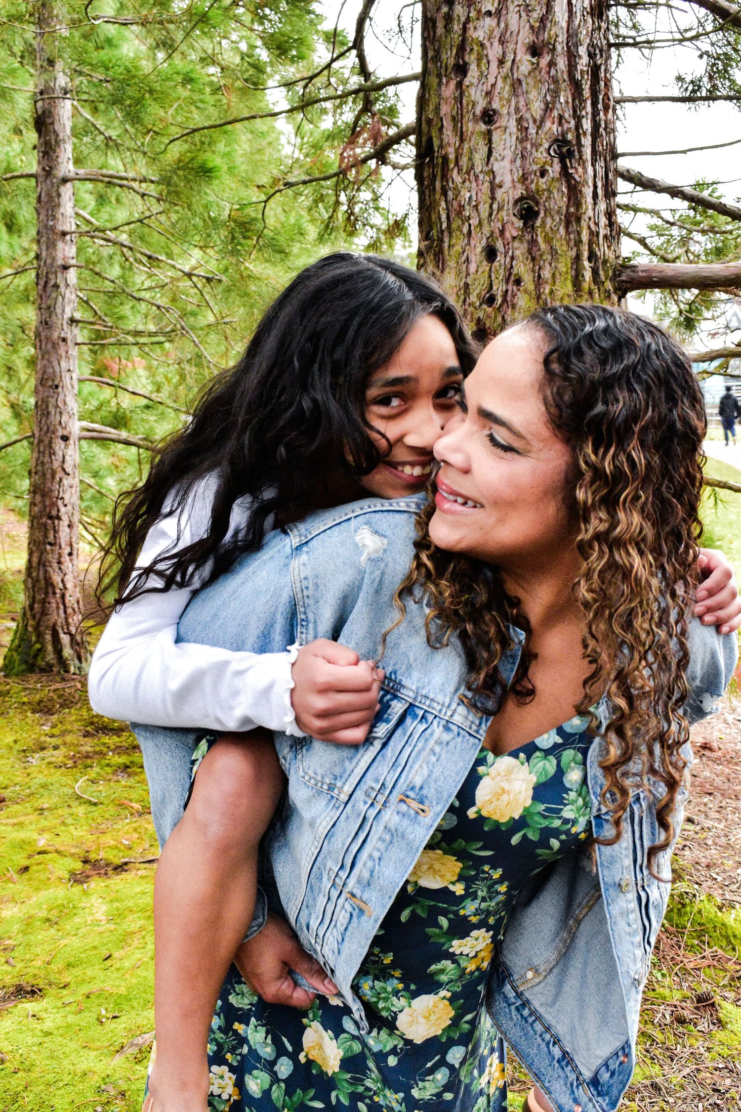 Mother and daughter sharing a joyful embrace while wearing denim jackets in a peaceful forest setting with green moss and trees. Seattle