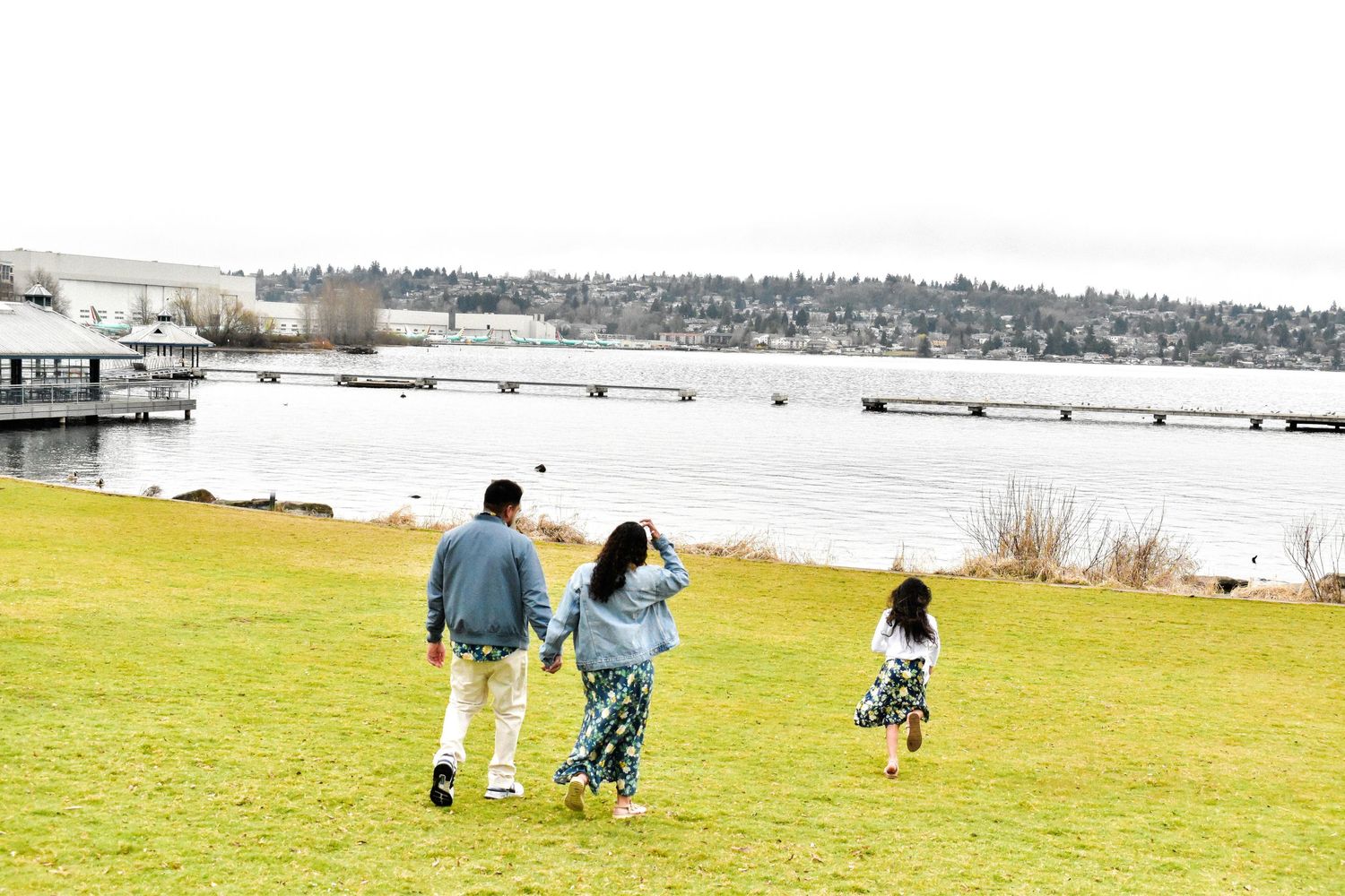 Seattle family photography. Family walking together on grassy slope overlooking waterfront with docks and boats on a cloudy day.