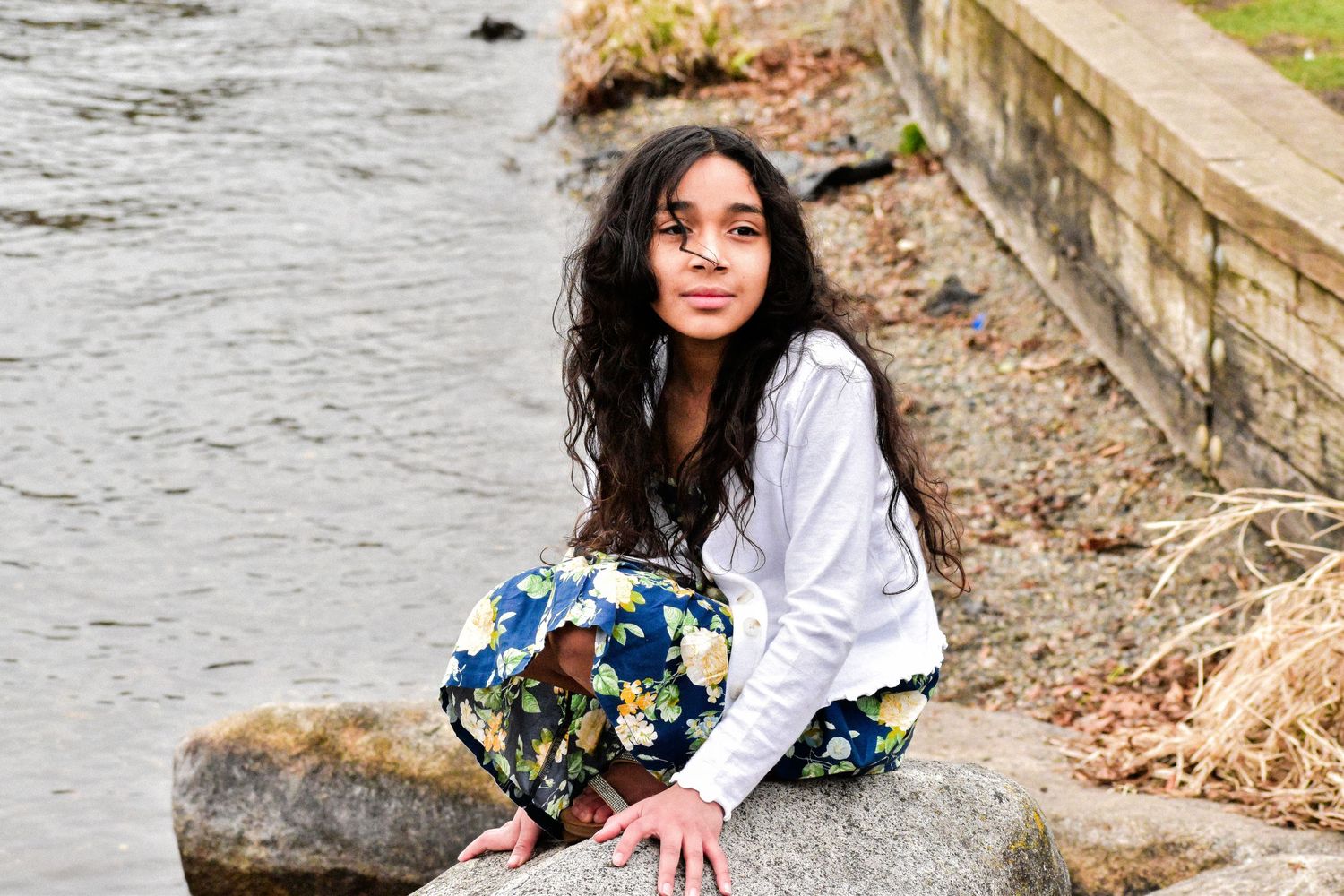 Seattle child photography. Someone sits casually on a large rock by a flowing river wearing a white shirt and floral pants on an autumn day.