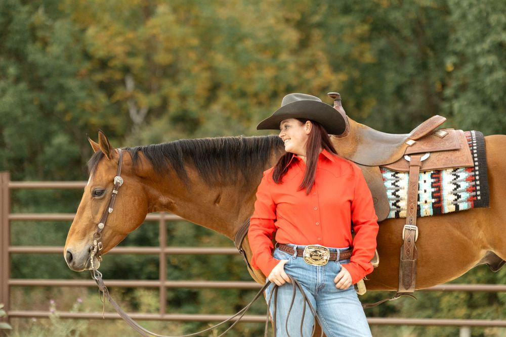 A brown horse with western saddle stands beside rider in red shirt and cowboy hat.
