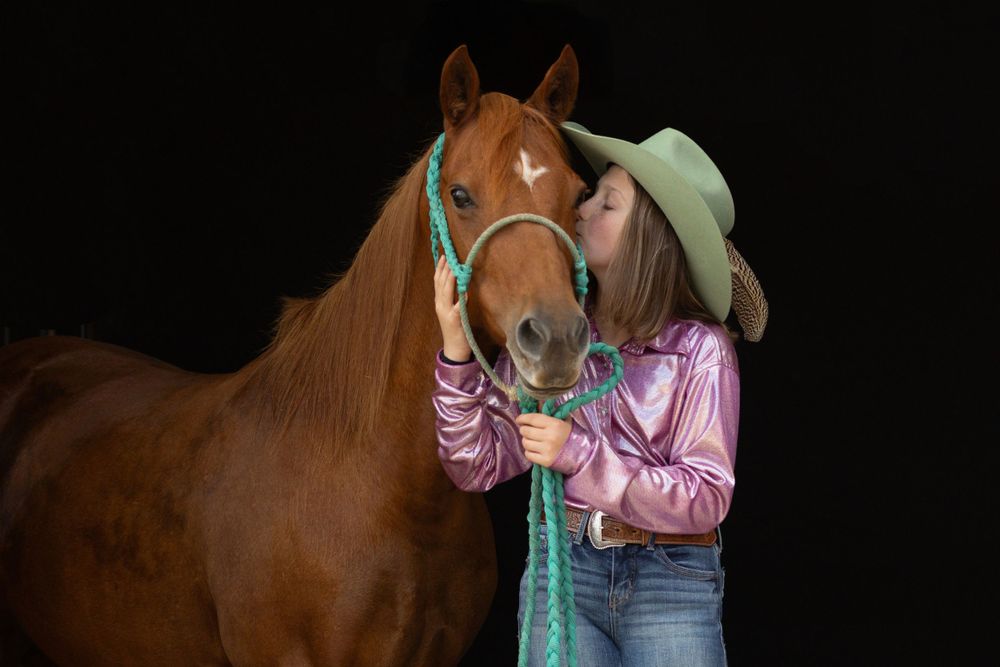 Gentle moment between horse and handler wearing pink shirt and mint green hat against dark background.