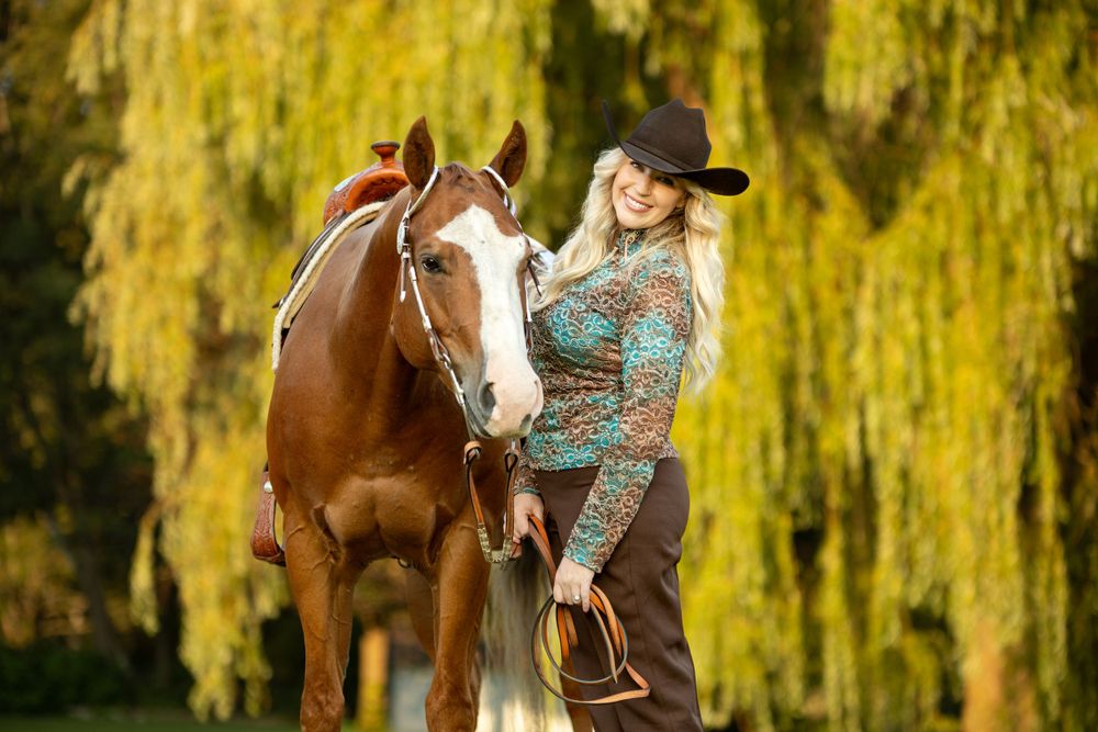 A brown horse with white markings stands with rider in western attire against yellow willow backdrop.