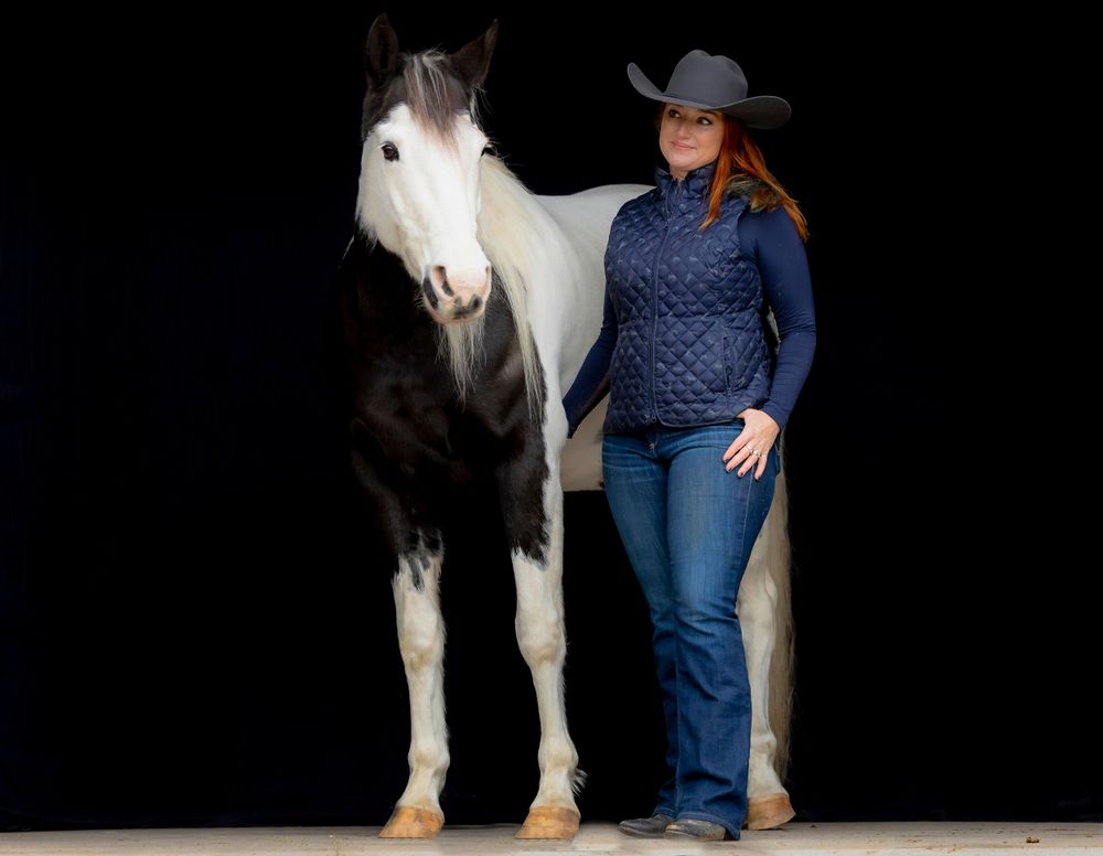 A black and white paint horse stands next to someone in western wear against a dark background.