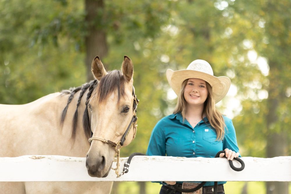 Horse and handler pose by white fence on sunny day with trees in background.