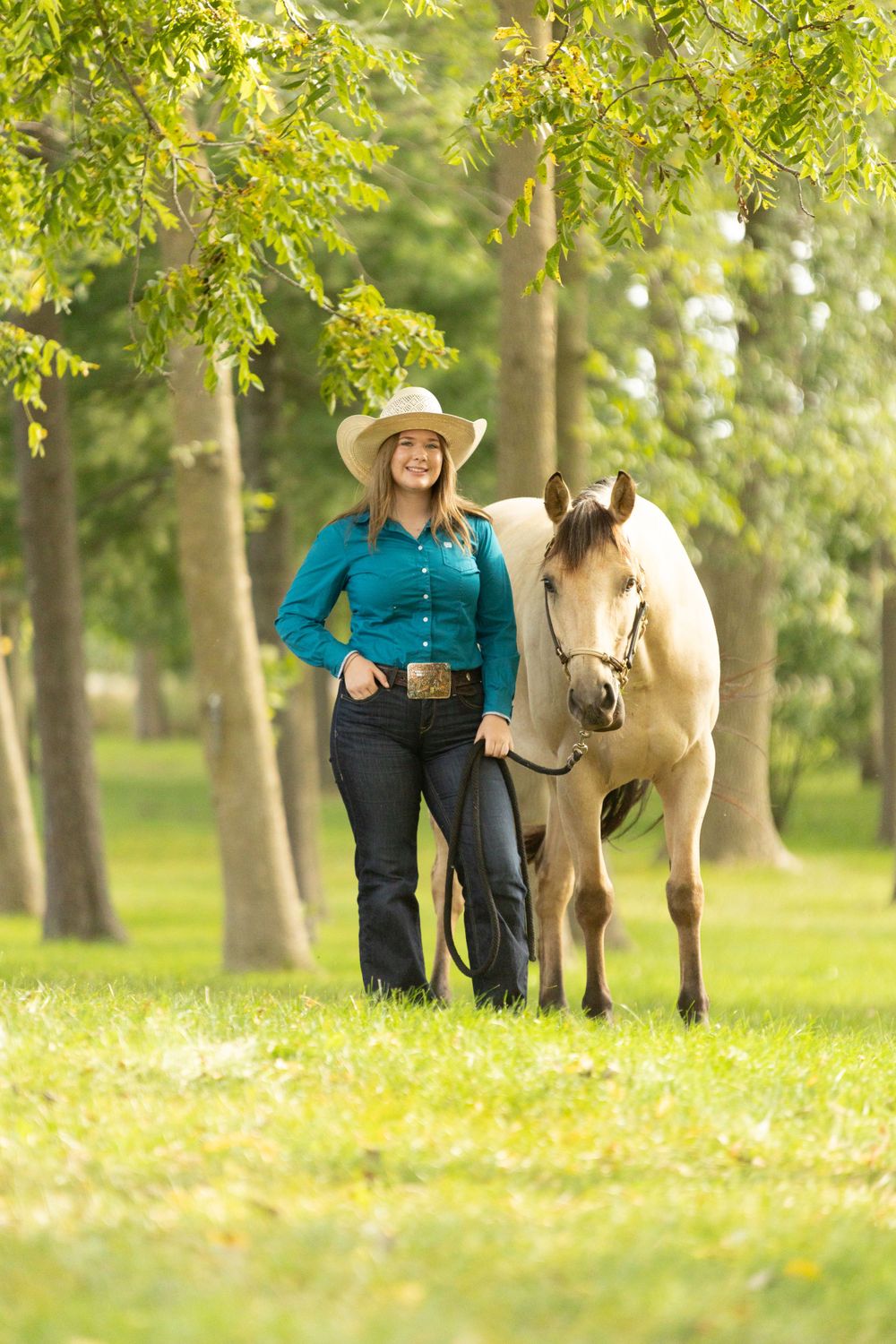 Horse and handler in turquoise shirt walk through sunlit park with trees.