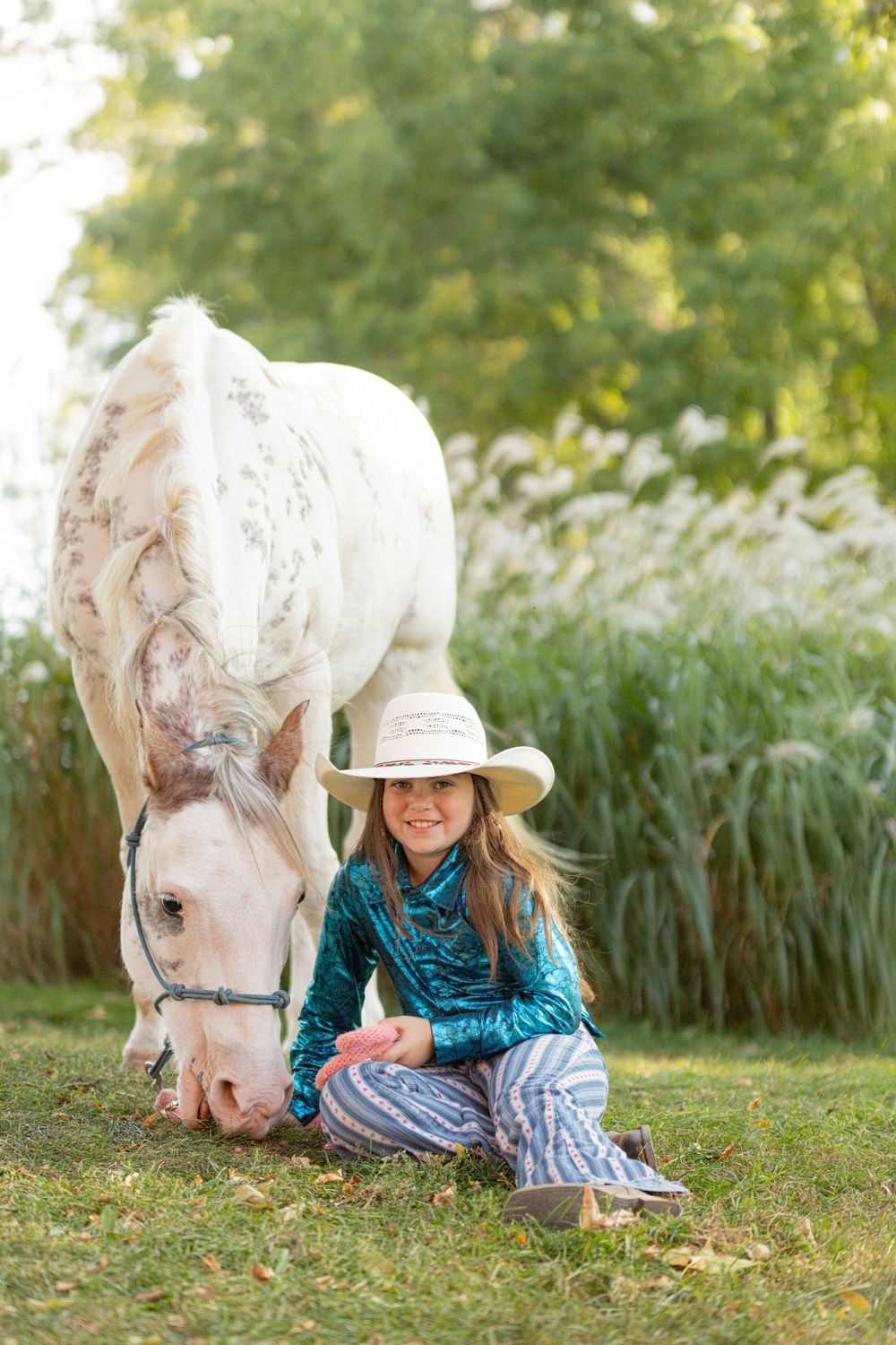 A white horse and rider in a turquoise top relax in a grassy field with ornamental grasses in the background.