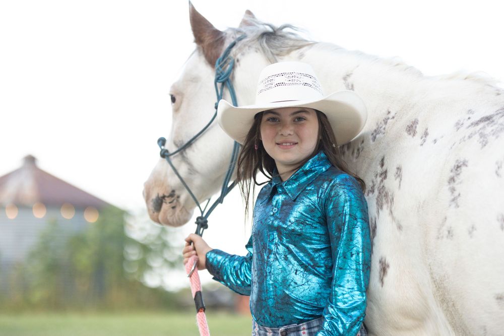 A white horse and rider in a sparkly turquoise jacket pose together on a sunny day.