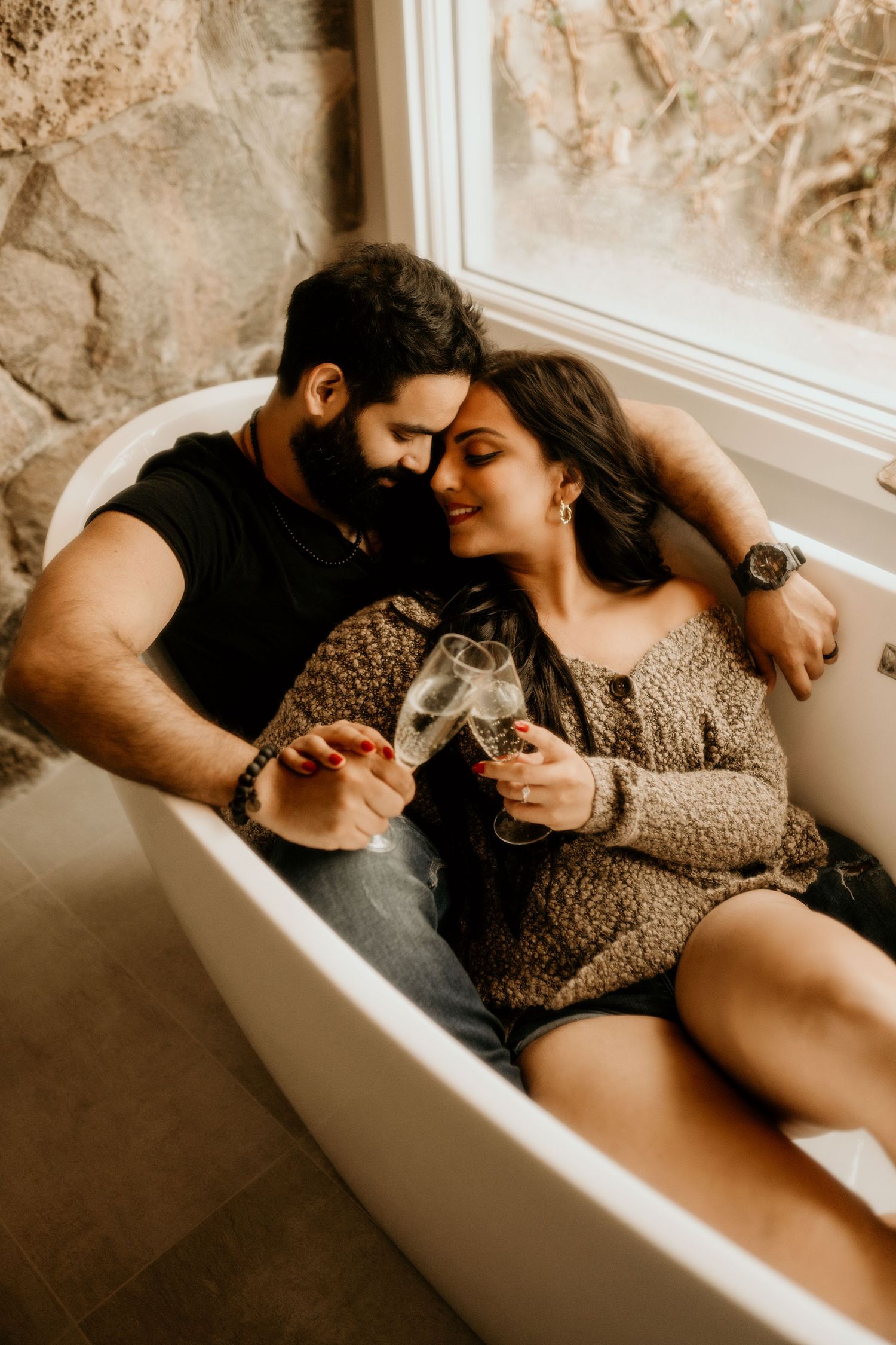 A couple shares an intimate moment while sitting in a vintage bathtub, holding beverages and dressed in cozy attire.