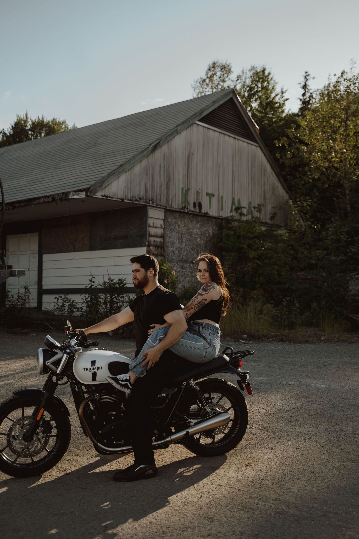 Two people ride a vintage motorcycle in front of a weathered barn at dusk.