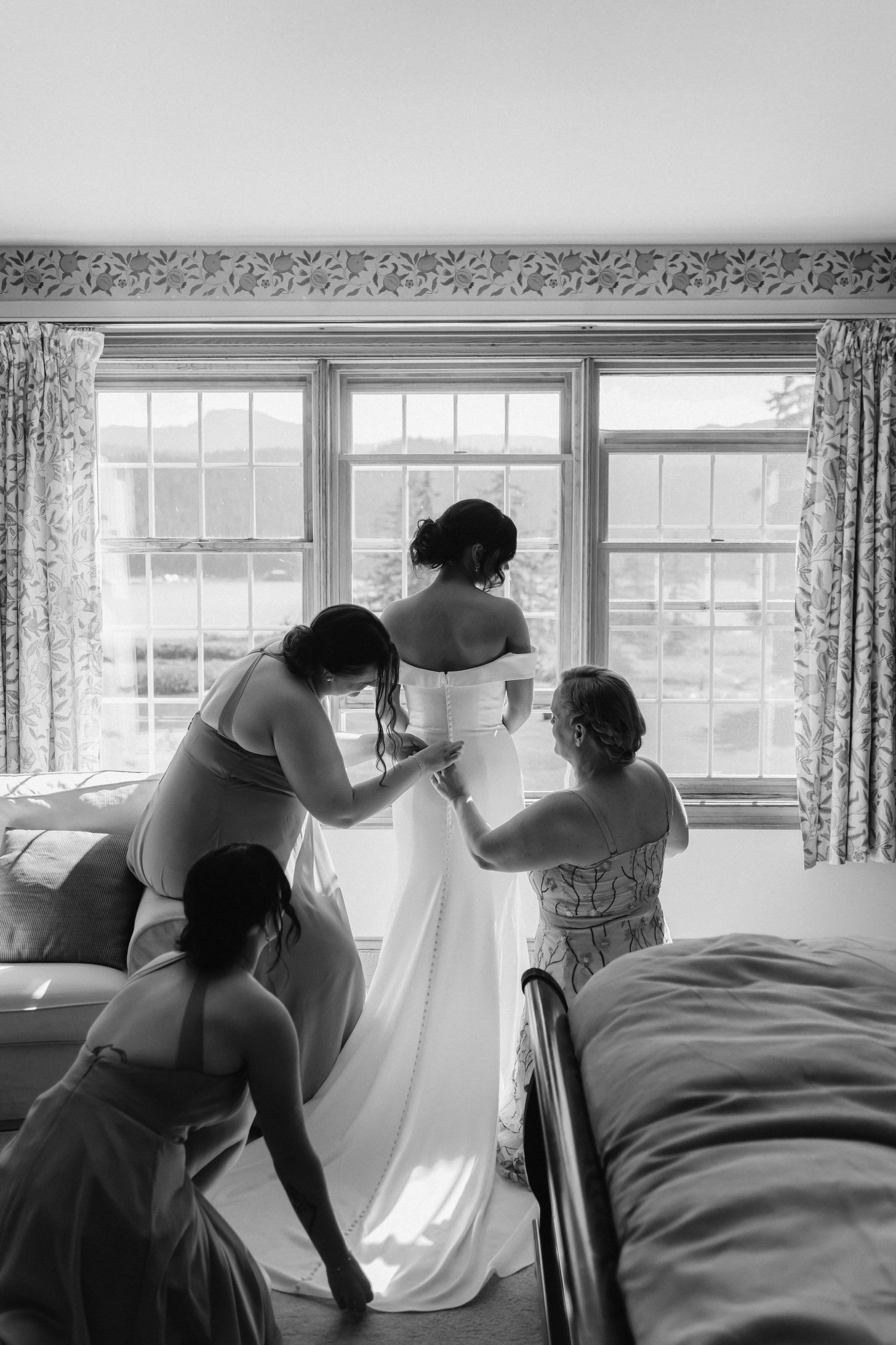 Bridal party assisting with wedding dress preparations by sunlit windows.