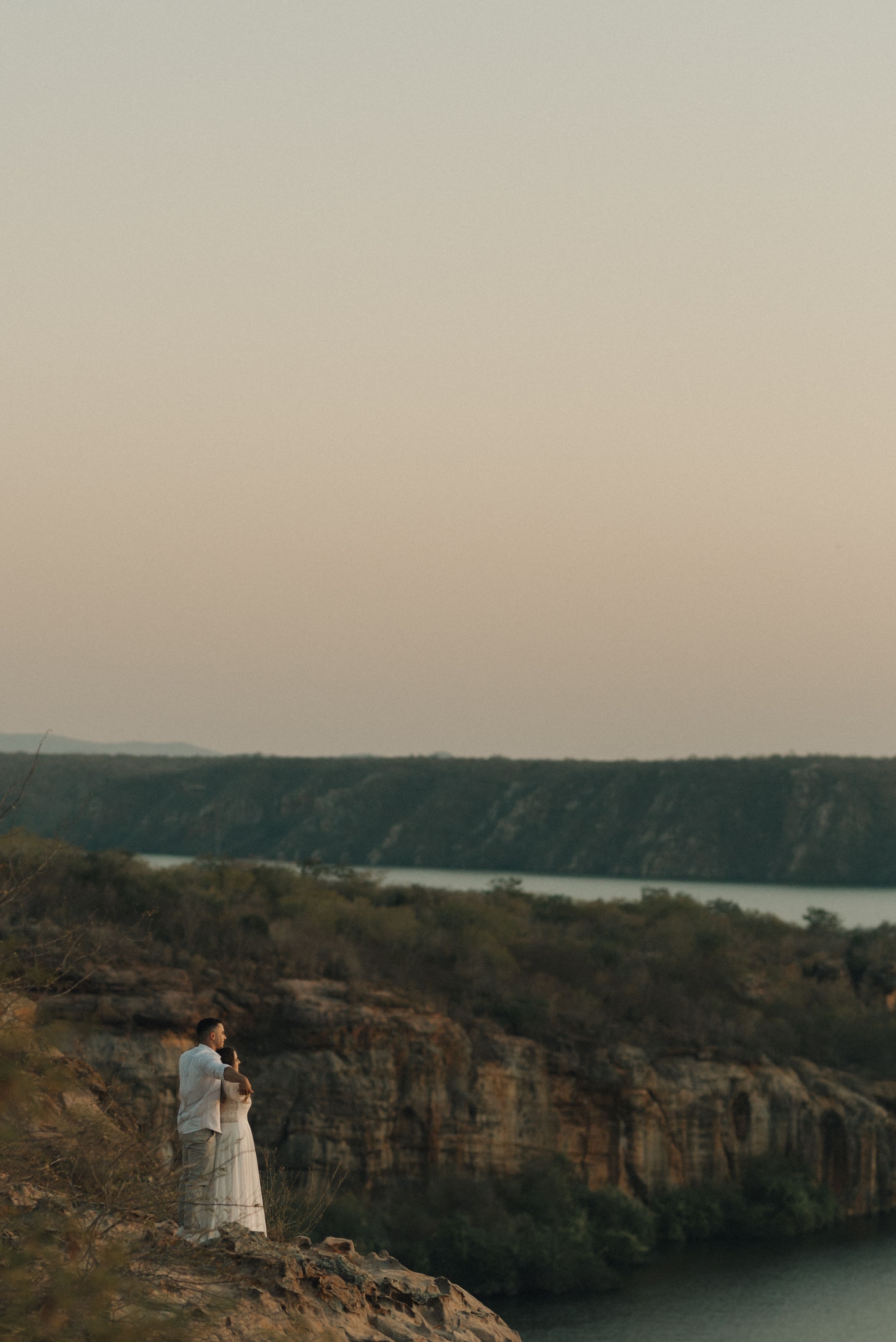 Couple in white outfits standing on cliff edge overlooking a scenic reservoir at sunset.