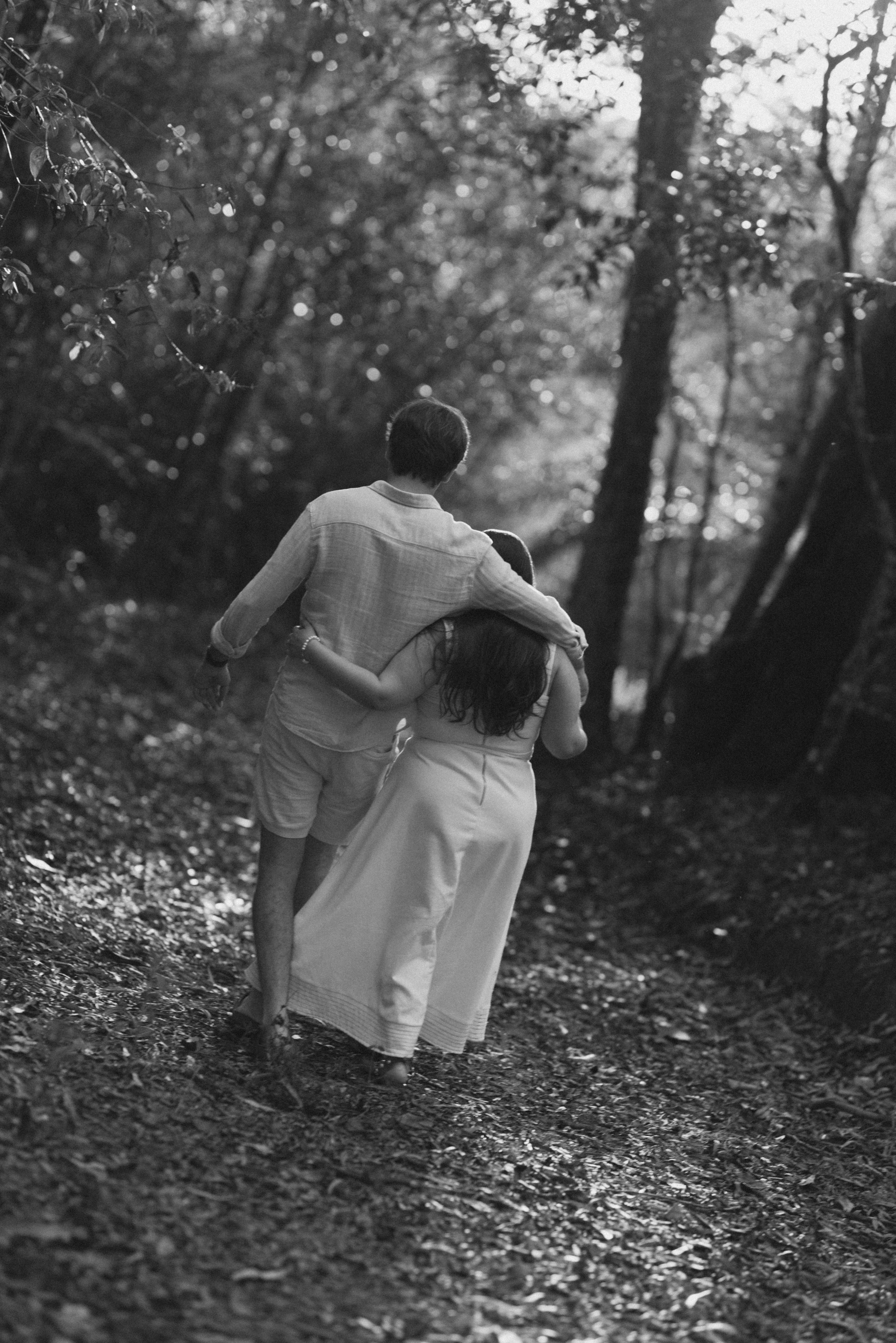 A couple in white attire walks together along a wooded forest path in black and white photography.