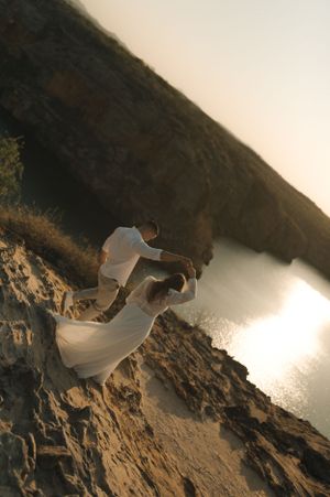 Couple in white attire standing on rocky cliff edge overlooking water in dramatic lighting.