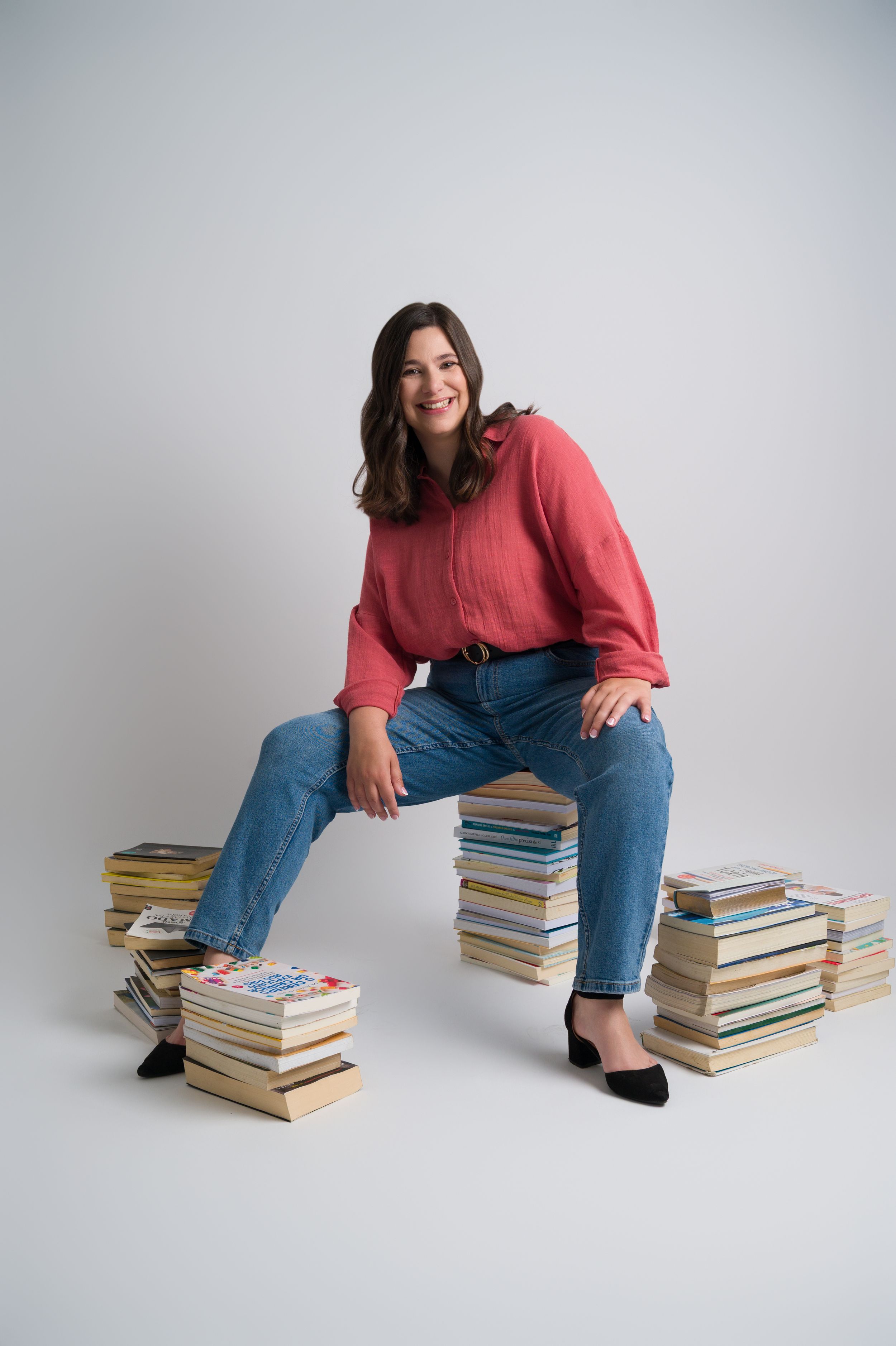 Cheerful woman in red shirt and jeans poses with stacks of books in studio.