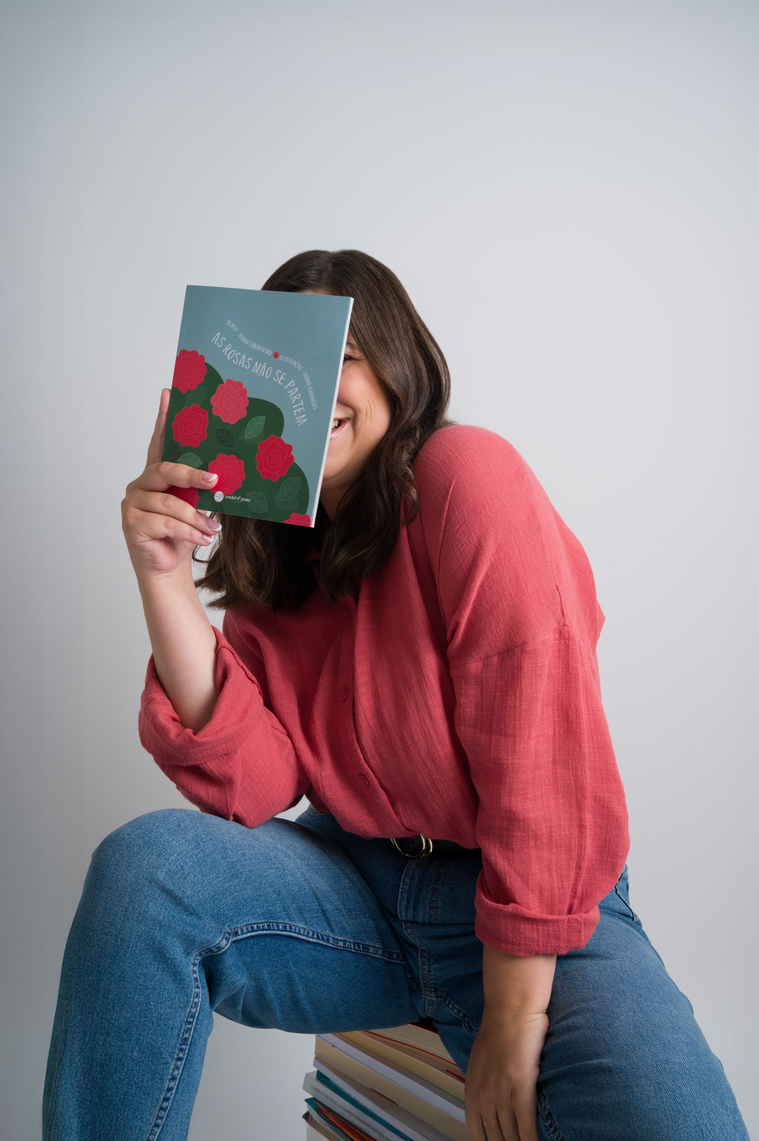 Woman in red shirt hides face behind colorful book while sitting on book stack.