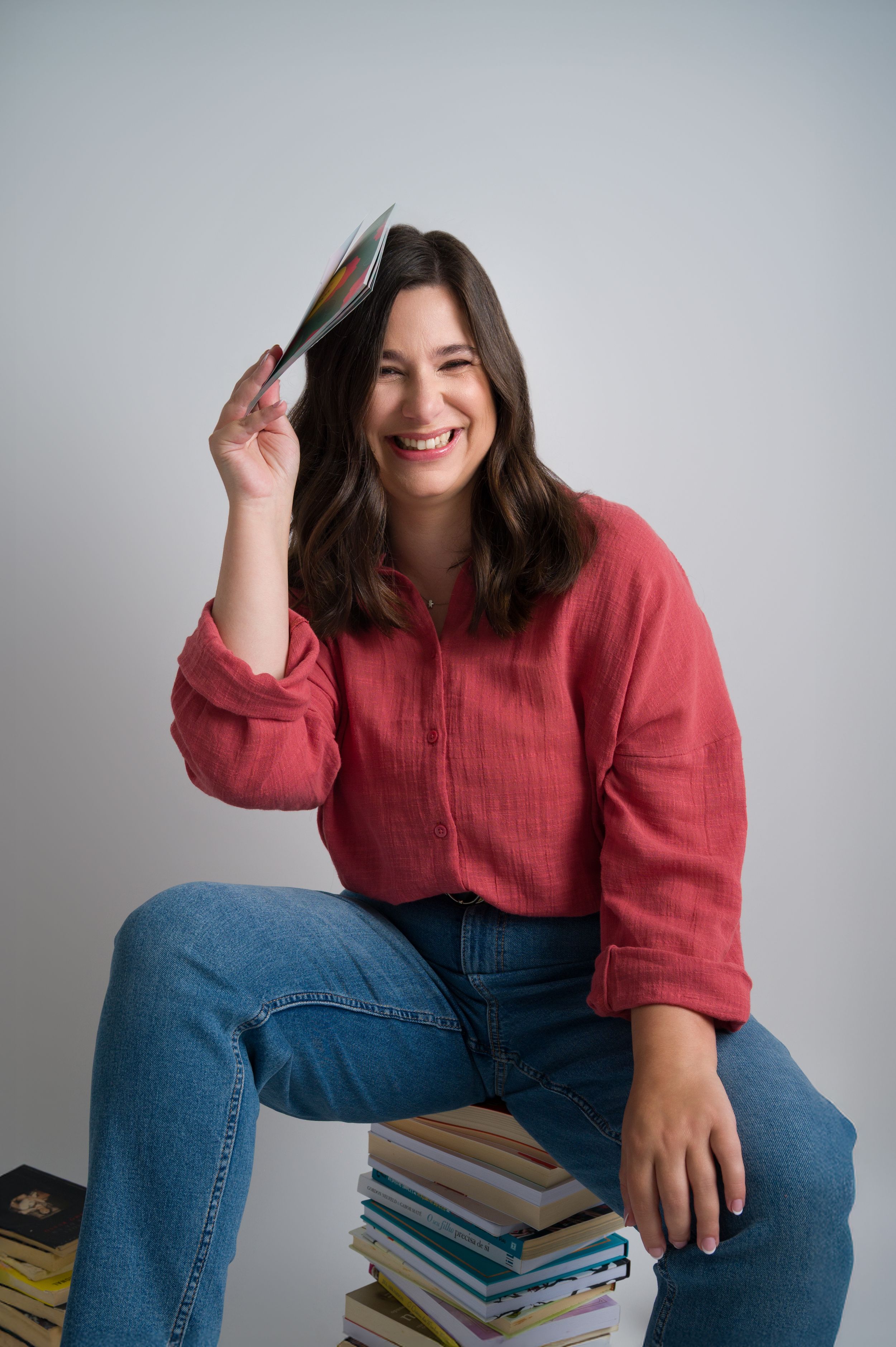 Smiling woman in red shirt sits on stack of books holding one above her head.