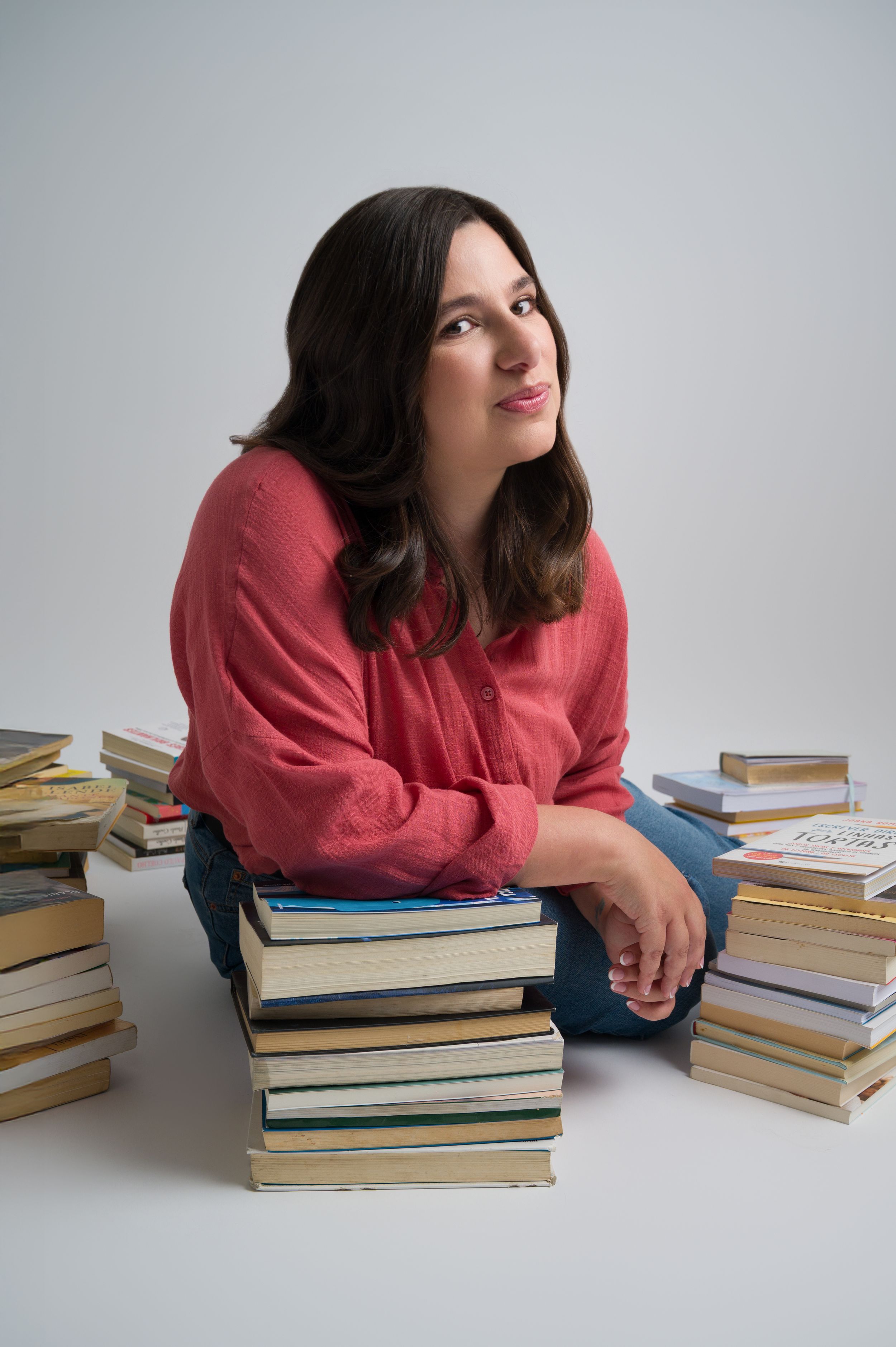 Woman in red blouse sits thoughtfully among stacks of books on white background.