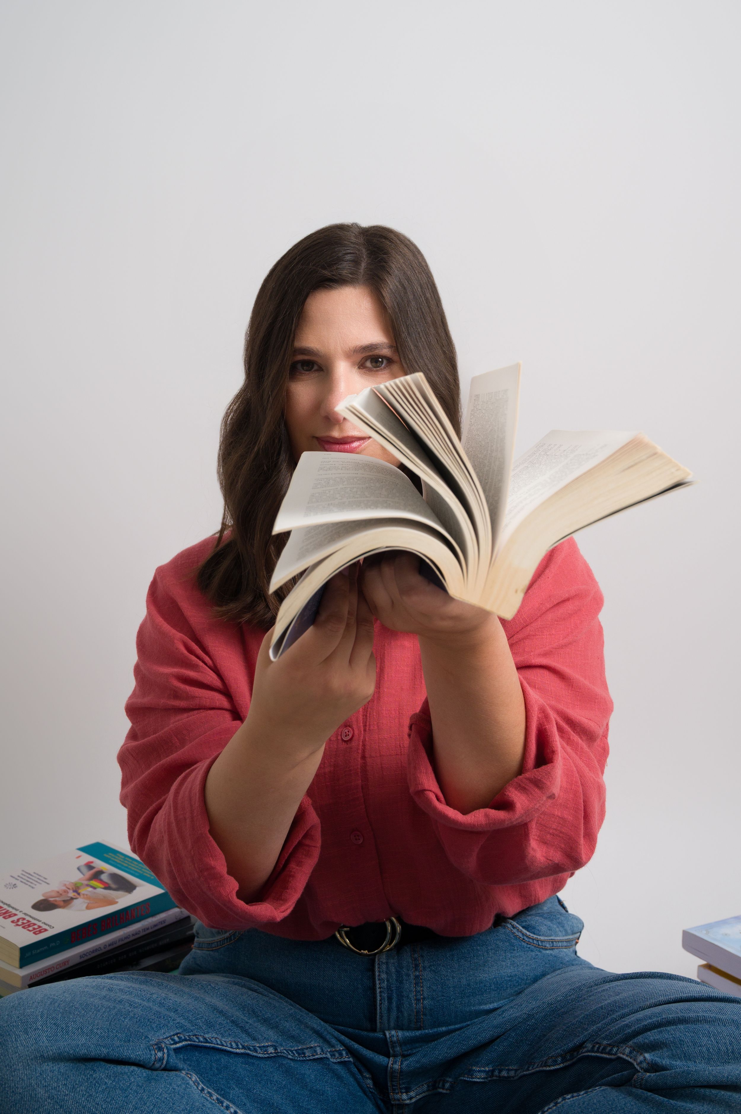 Woman in red shirt fans open book pages toward camera surrounded by books.