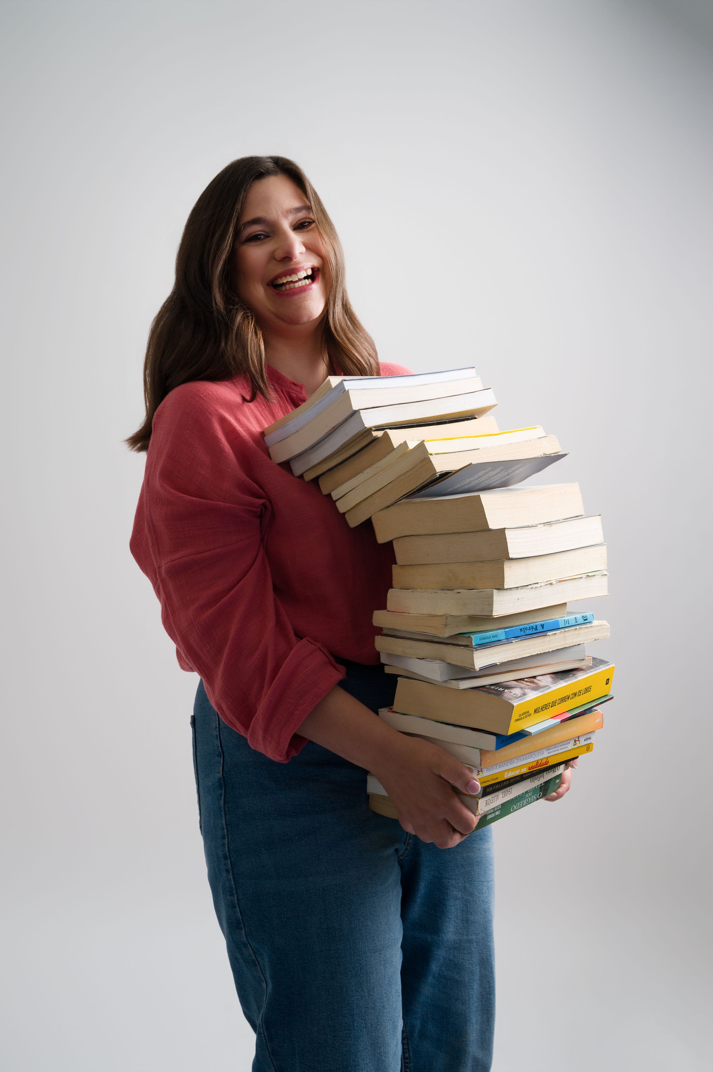 Laughing woman in red shirt carries tall wobbling stack of books in studio.
