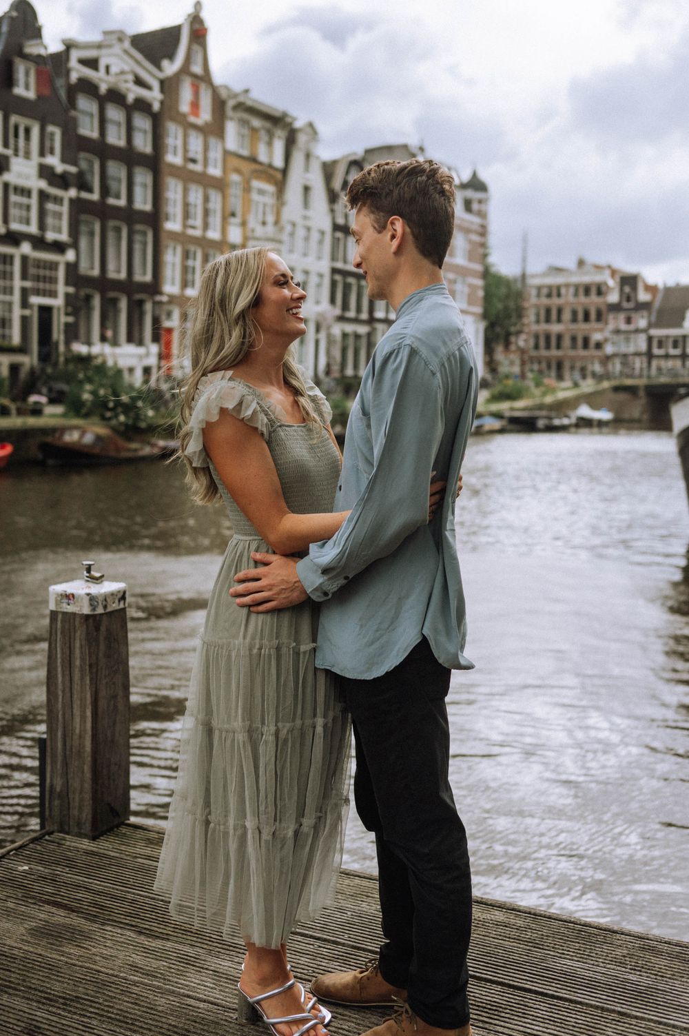 A couple embraces on a wooden dock with traditional Amsterdam canal houses in the background.