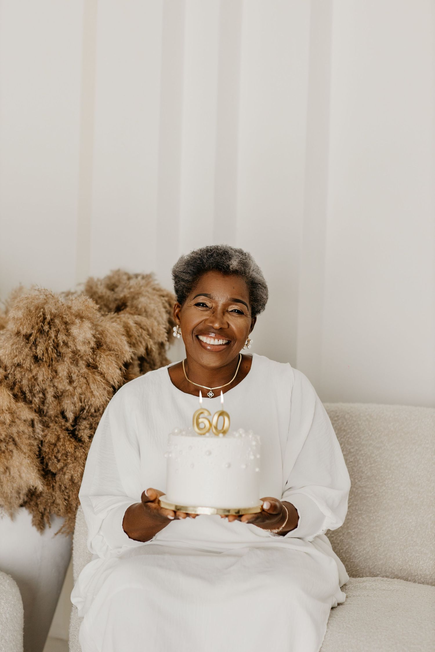 Woman in white dress holding a birthday cake with number 60 candles sitting on couch next to pampas grass decor.
