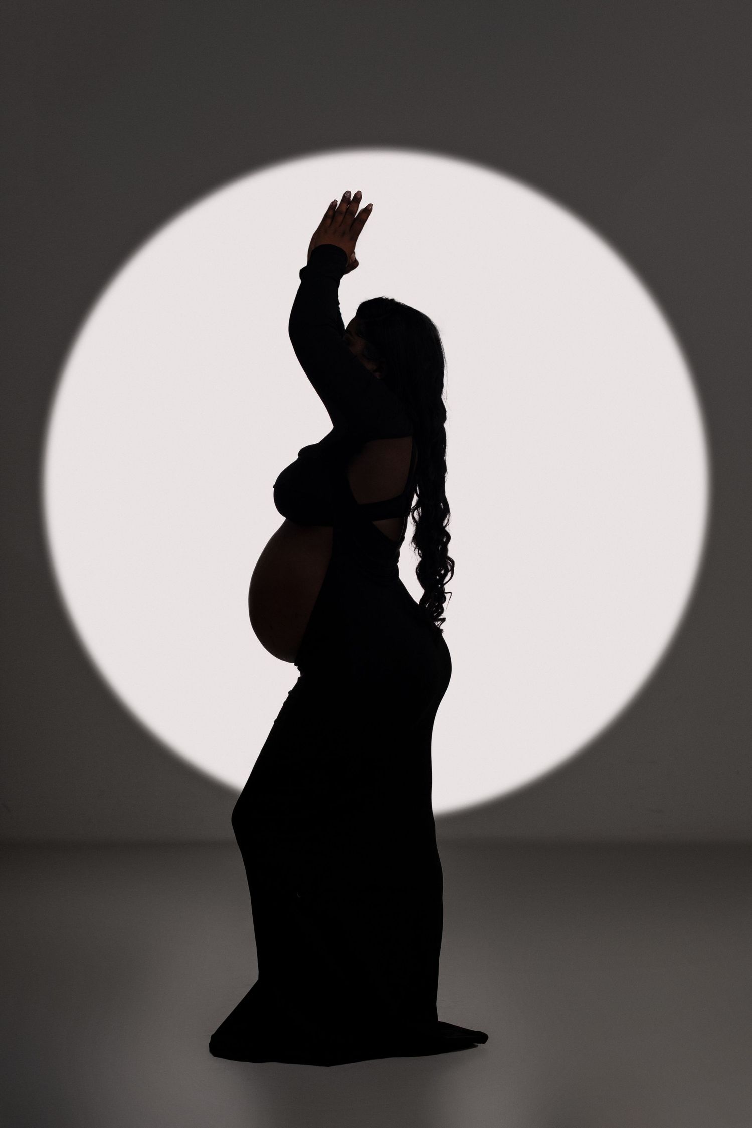 Artistic silhouette of pregnant woman in long dress against white circular backdrop.