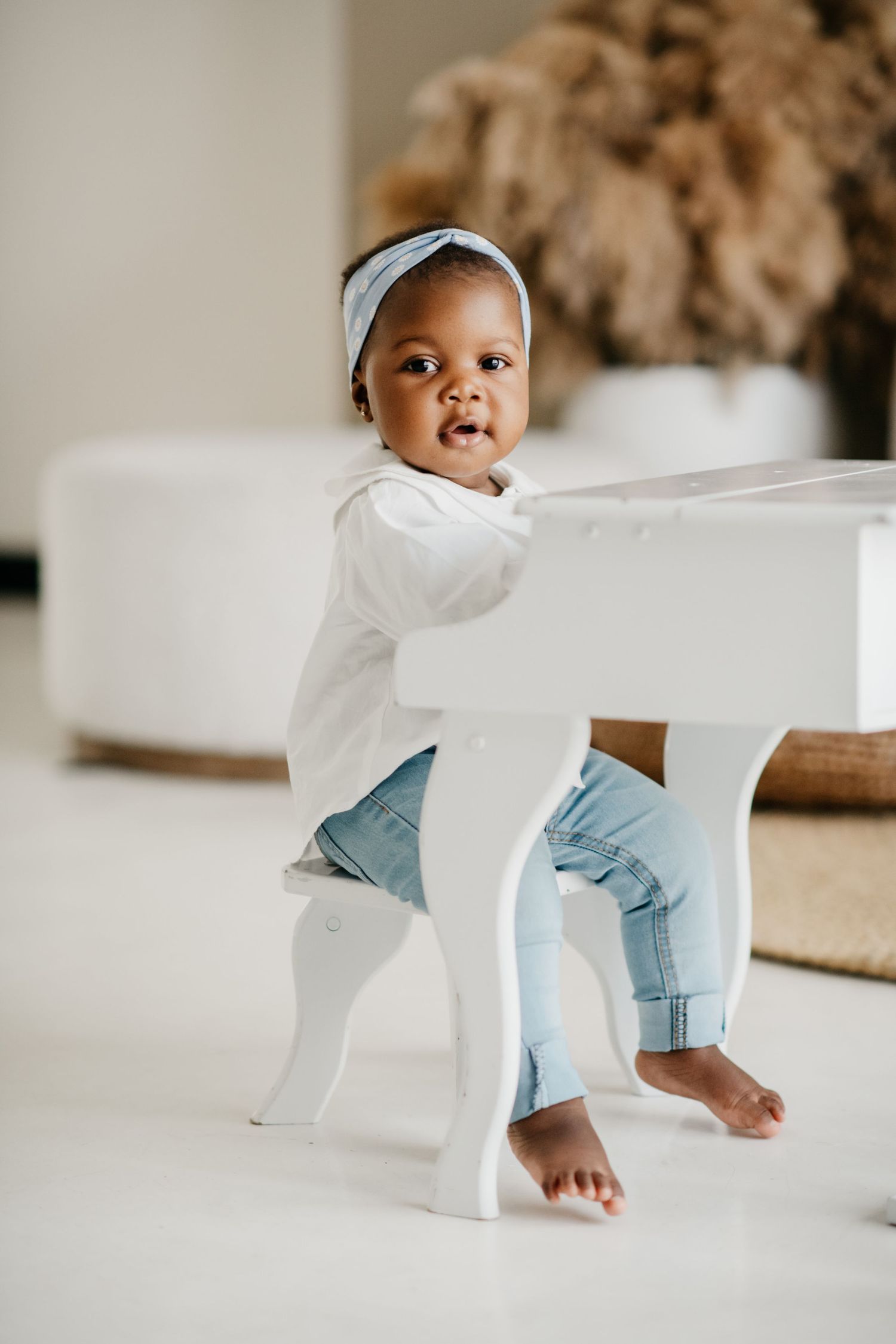 Toddler in white shirt and blue jeans peeking behind white chair with pampas grass decor.