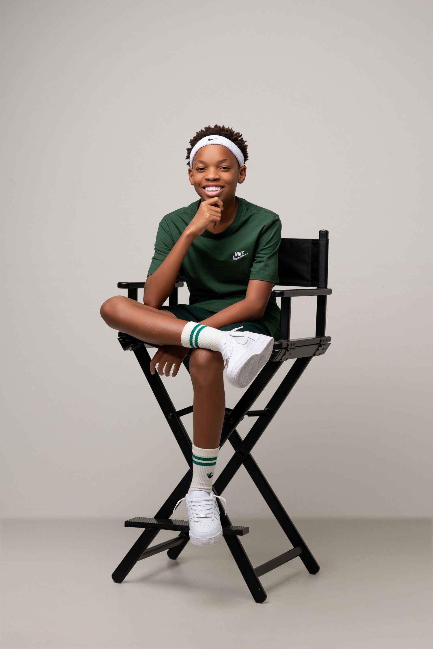 Young person in green Nike shirt and white shorts sitting casually in director's chair.